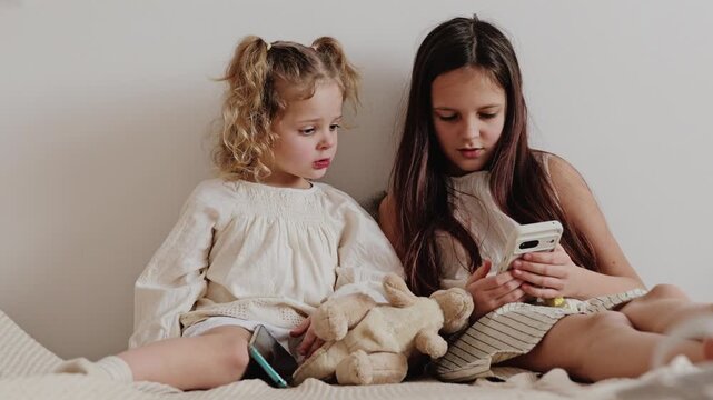 Two children sit together on a bed, one girl is using a smartphone while the other looks on and holds a stuffed animal in a simple room