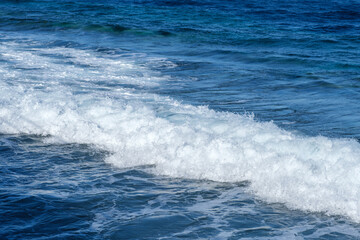 Ocean waves crashing onto the shore, creating foam and ripples against a backdrop of deep blue water and clear sky
