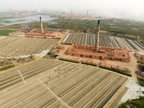 Aerial view of rows of bricks under the sun next to brick kilns with tall chimneys casting shadows on the ground, Aminbazar, Dhaka Division, Bangladesh.