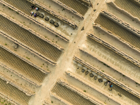 Aerial view of workers meticulously arranging dark, circular objects within the sandy, geometric patterns of a brickyard, Aminbazar, Dhaka Division, Bangladesh.
