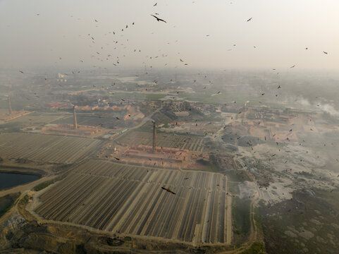 Aerial view of brick kilns punctuate the landscape, smoke billowing amidst a swarm of birds in a hazy scene, Aminbazar, Dhaka Division, Bangladesh.