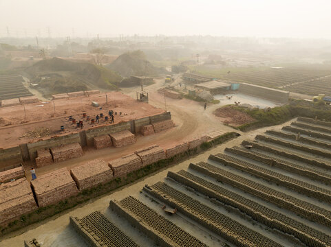 Aerial view of brick stacks under a hazy sky with workers, creating a textured landscape of earthy tones and industrial activity, Aminbazar, Dhaka Division, Bangladesh.