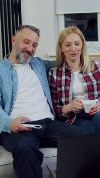 Family leisure concept where good-looking in high spirits smiling blond woman and her bearded husband relaxing on comfortable sofa at home and revisioning interesting video on computer