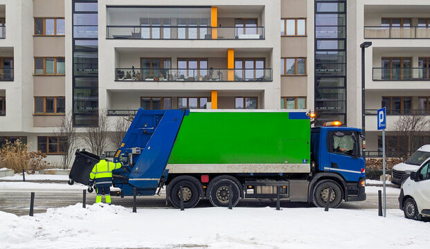 Blue cab and green container garbage truck with sanitation worker standing near rear section on city street with apartment buildings.