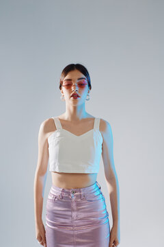 Young woman in a white crop top and shiny skirt poses confidently wearing pink sunglasses. Studio shot with soft lighting. Concept of woman beauty fashion and style.