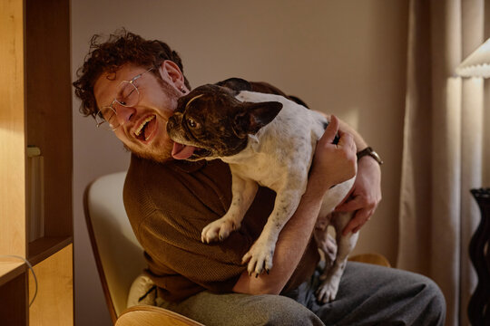 Caucasian young adult man sitting and hugging French Bulldog, smiling with eyes closed while dog licking his face, showing joyful interaction between pet and owner in home setting