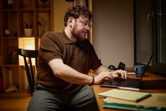 Caucasian young adult man sitting at desk typing on laptop, wearing glasses and beard, working late at home office, focused on screen with notepad and pen on table