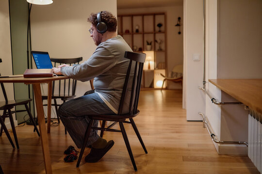 Caucasian man wearing headphones working on laptop at wooden table in modern home interior, sitting on chair with focused expression, bookshelf and open doorway in background
