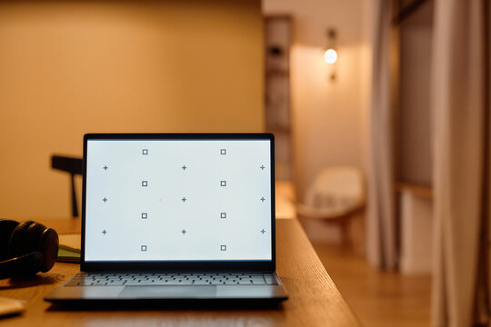 Laptop with blank screen displaying calibration marks standing on wooden desk in modern home interior, headphones lying nearby, blurred background with soft lighting