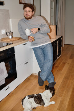 Caucasian young adult man with curly hair and beard leaning against kitchen counter holding mug and cookie, wearing glasses and slippers, French Bulldog lying on wooden floor nearby