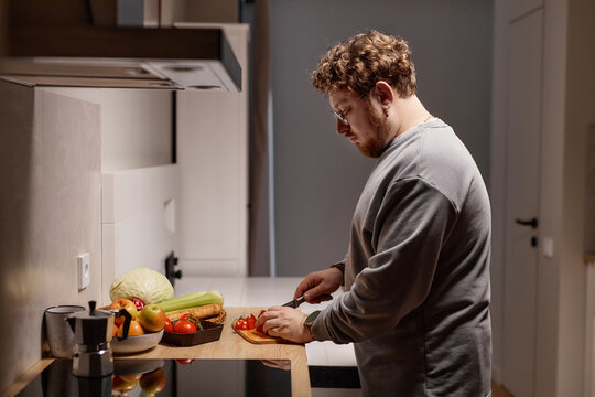 Caucasian young adult man preparing meal by slicing fresh vegetables on kitchen counter, focusing on cutting tomato with knife, surrounded by various produce and kitchenware