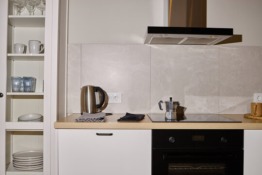 Modern kitchen countertop featuring electric kettle, coffee maker, cup and saucer, with glassware and plates arranged in cabinet, showcasing clean and organized cooking space