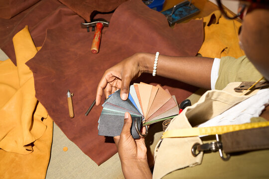 Black woman selecting leather color swatches in workshop, holding fan of material samples over brown and tan leather hides, tools and measuring tape visible on work surface