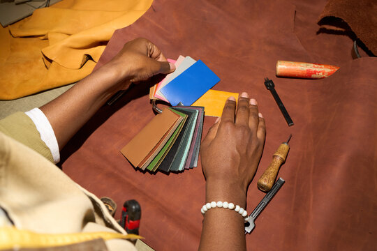 Black young adult woman selecting leather color swatches with both hands on brown leather hide, holding tool set including awl and pricking iron on workshop table, wearing bracelet