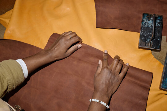 Black woman hands working with brown leather on workshop table, carefully aligning material for crafting project, visible tools and yellow leather pieces surrounding workspace