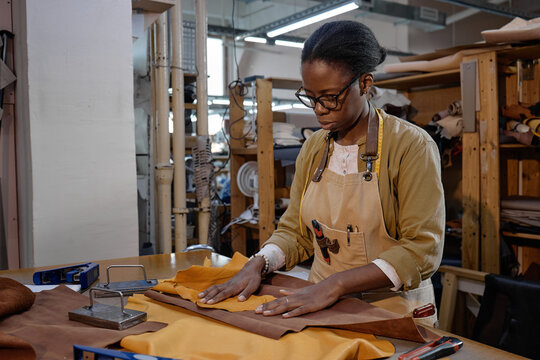 Black woman young adult working in leather workshop cutting brown leather fabric on worktable using metal ruler and tools, focused on precise craftsmanship surrounded by materials and equipment