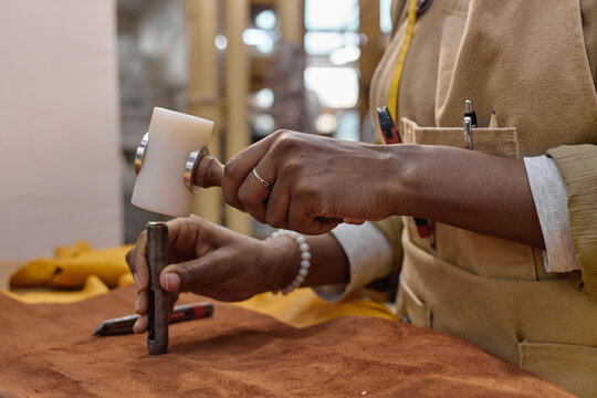 Black woman young adult working in leather workshop using mallet and chisel shaping leather piece, hands visible holding tools, focus on craftsmanship and manual technique