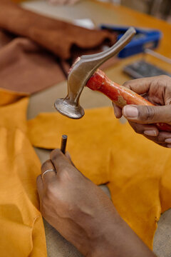 Black woman hands working with leather using hammer and metal punch tool in workshop, creating precise hole in tan leather material, demonstrating craftsmanship and manual skill