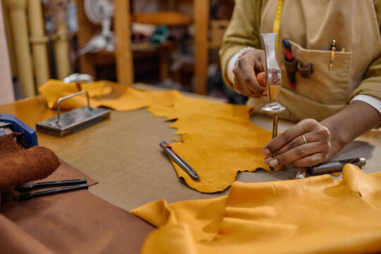 Black man working in leather workshop using specialized tool to cut leather hide, hands visible holding tool and measuring tape draped around neck, crafting handmade leather goods