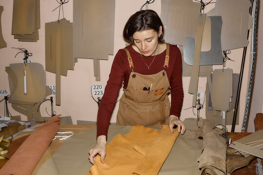 Young adult Caucasian woman working in leather workshop inspecting large piece of tan leather on worktable surrounded by leather patterns and tools, focused on craftsmanship process