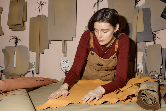 Caucasian young adult woman working in leather workshop, carefully inspecting and smoothing large piece of leather on worktable, surrounded by leather patterns and material rolls