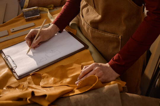Caucasian woman sketching leather pattern on clipboard while working in workshop, left hand holding piece of leather, measuring tape and crafting tools visible on table