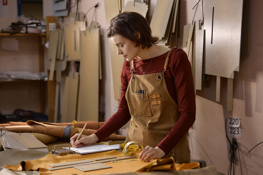 Young adult Caucasian woman working in leather workshop sketching design on paper with pencil, surrounded by leather pieces and measuring tape, focusing on creative process