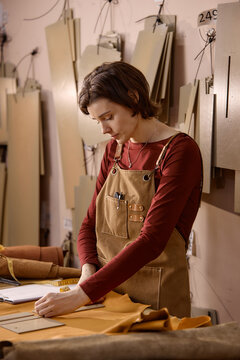Young adult Caucasian woman working in leather workshop, measuring and cutting leather material on worktable, focused on craftsmanship, surrounded by leather pieces and tools