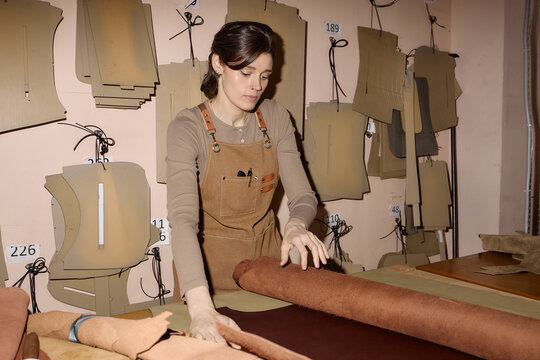 Young adult Caucasian woman working in leather workshop handling large roll of brown leather, surrounded by hanging leather patterns and tools, focused on preparing materials for crafting