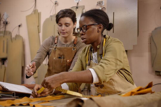 Young adult Caucasian woman and young adult Black woman working together in leather workshop, measuring leather material with tape measure, discussing project, wearing aprons, focused on craftsmanship
