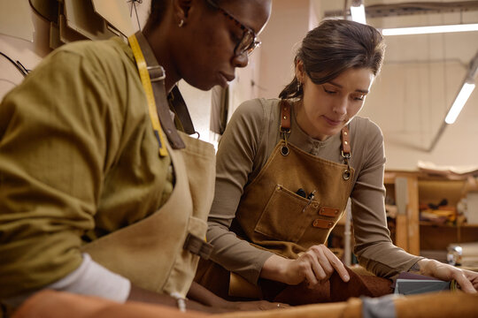 Caucasian young adult woman and Black young adult woman working together crafting leather goods in workshop, both wearing aprons and focusing on detailed handwork at workbench