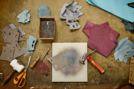 Leather pieces, metal rivets, scissors, hammer, awl, and crafting tools arranged on workbench, showing process of handcrafting leather goods in artisan workshop setting