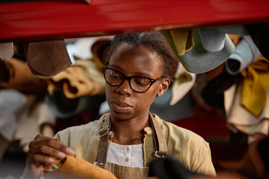 Black young adult woman wearing glasses working in leather workshop inspecting piece of leather material surrounded by various leather hides hanging in background