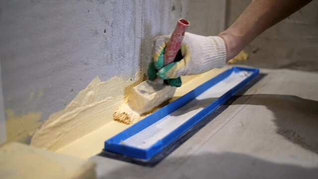 Close-up of a worker applying yellow liquid waterproofing with a brush around a shower drain protected with blue tape. Bathroom renovation process before tile installation.