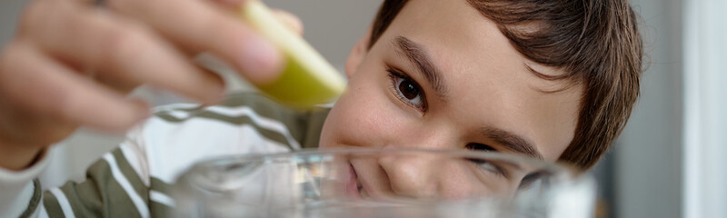 Naklejka premium Portrait of boy smiling while holding green apple slice above glass bowl, focusing on fruit with joyful expression, close up of child engaging in healthy eating activity