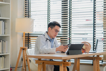 Asian male doctor using smartphone while sitting at medical office desk with tablet laptop and anatomy brain model professional healthcare consultation concept modern clinic workplace environment.