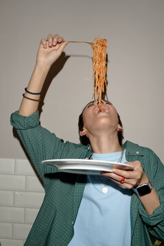 Teenage boy holding plate while lifting spaghetti above mouth, tilting head back and preparing to eat, wearing casual clothing and smartwatch, standing indoors