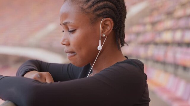young woman in stadium leaning on railing with head down, earphones in, exhausted and reflective closeup of athlete catching breath, empty bleachers background, quiet healing mood