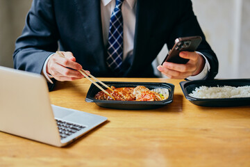 Asian businessman eating bento while checking smartphone at office desk (face not visible)