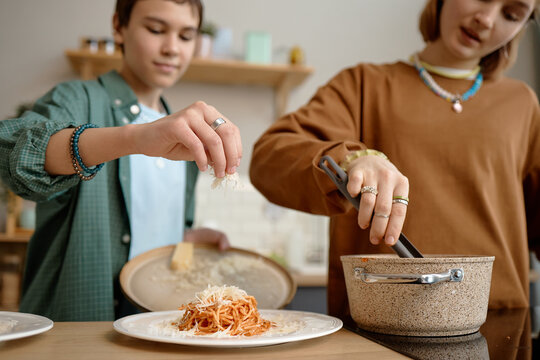 Teenage boy sprinkling grated cheese onto plate of spaghetti while teenage girl stirring sauce in pot, both preparing meal together in kitchen setting