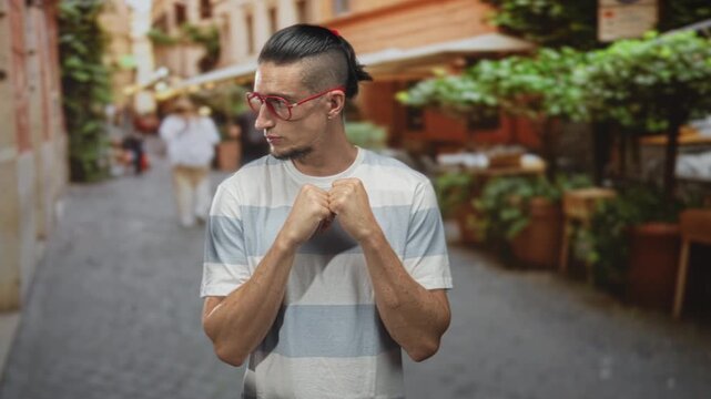 Man with fists loosely clasped at chest wearing red glasses on a cobbled street with outdoor cafe seating and potted plants; quiet contemplation.
