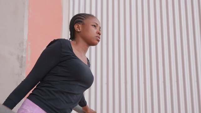 black woman walking past corrugated wall, side profile showing cornrows and athletic top, pastel concrete backdrop creating minimalistic urban scene, focused stride and determined expression, commuter