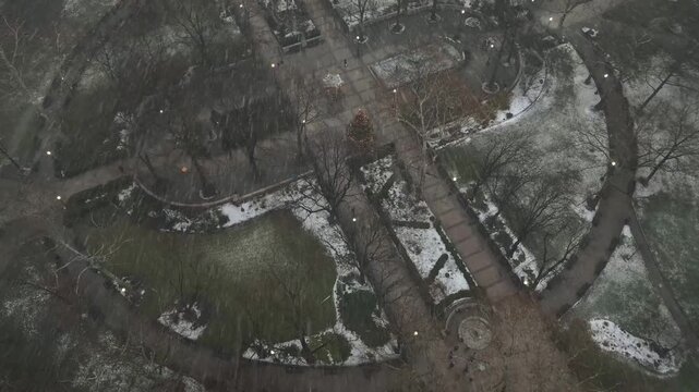 Rittenhouse Square in Philadelphia during light snowfall, a decorated Christmas tree at the center as snow dusts pathways and bare winter trees. Aerial top down landing.