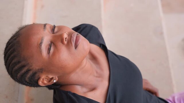 black woman with cornrows sitting on concrete stairs, wearing black top and purple pants, relaxed pose and upward gaze, soft daylight and textured steps, close aerial portrait capturing calm