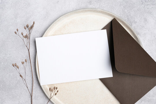 Wedding invitation card mockup with blank white space on a round plate, accompanied by a dark brown envelope and dried flowers for decorative elements