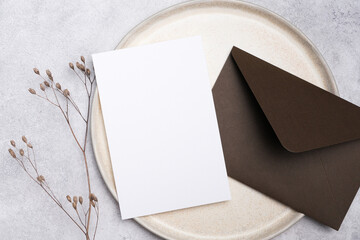 Wedding invitation card mockup with blank white space on a round plate, accompanied by a brown envelope and dried flowers on a textured gray background