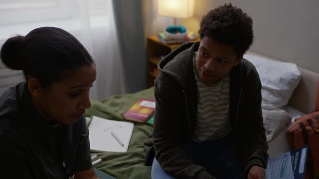 Slowmo shot of young Black female social worker and teenage boy holding social assistance brochures and discussing scholarship for underprivileged students sitting on bed surrounded by textbooks