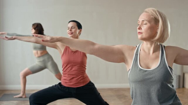 Three women doing Virabhadrasana II pose, their legs spread wide apart, one knee is bent, and their arms stretched out to sides at shoulder level during yoga class