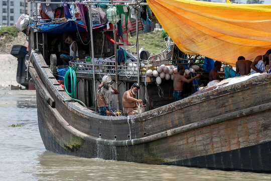 Chattogram, Bangladesh - 15 August 2025: View of men aboard a weathered wooden boat, its hull streaked with moisture, under a bright yellow tarp, amidst the muddy waters near the shore.
