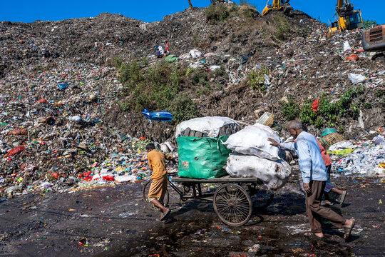 Chattogram, Bangladesh - 26 November 2025: View of laborers pushing a cart laden with refuse against a backdrop of a vast, towering landfill under a clear sky.
